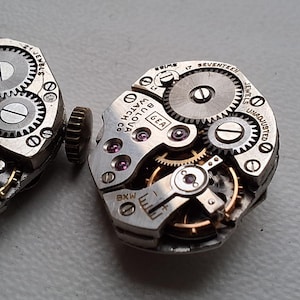 Peut inclure: Deux mouvements de montre Bulova vintage, avec des engrenages complexes, des bijoux et des composants m&eacute;talliques. Les mouvements de montre sont de couleur argent&eacute;e avec des accents dor&eacute;s, r&eacute;v&eacute;lant la m&eacute;canique interne des montres. Du texte est visible.