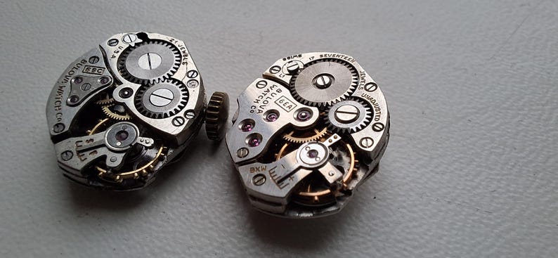 Peut inclure: Deux mouvements de montre Bulova vintage, avec des engrenages complexes, des bijoux et des composants m&eacute;talliques. Les mouvements de montre sont de couleur argent&eacute;e avec des accents dor&eacute;s, r&eacute;v&eacute;lant la m&eacute;canique interne des montres. Du texte est visible.