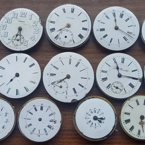 May include: A collection of antique watch faces with white dials and black Roman numeral hour markers. Some faces have sub-dials and delicate hands. The watch faces are arranged on a wooden surface, showcasing their aged appearance.