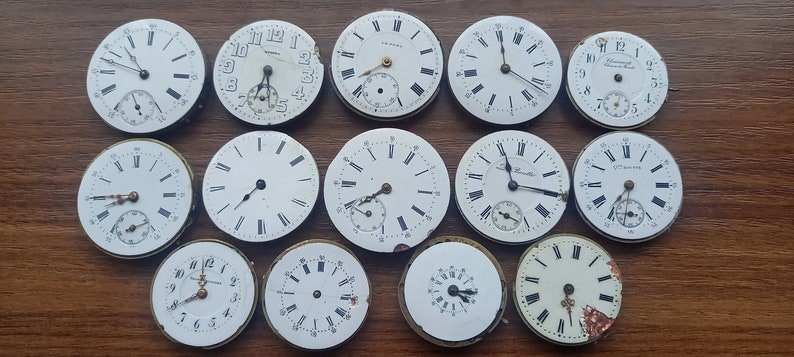 May include: A collection of antique watch faces with white dials and black Roman numeral hour markers. Some faces have sub-dials and delicate hands. The watch faces are arranged on a wooden surface, showcasing their aged appearance.