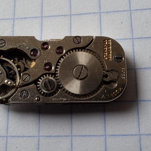 May include: Close-up of a vintage watch movement with intricate gears, screws, and jewels. The silver-toned mechanism is rectangular, with visible text and a circular balance wheel. The watch movement is a detailed example of horological engineering.
