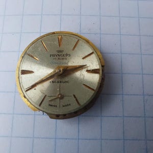 Könnte beinhalten: Vintage Pryngeps Zifferblatt mit goldfarbener Lünette und silbernem Zifferblatt. Das Zifferblatt hat goldfarbene Zeiger und Markierungen mit dem Text "PRYNGEPS 17 RUBIS Special INCARLOC SWISS MADE". Das Zifferblatt ist rund.