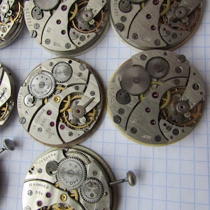 May include: Close-up of six antique watch movements with intricate gears and markings. The watch movements are silver and gold in color and have various numbers and text on them, including "15 KAMHEH", "2-53", "SU", "3602", "953393", and "237731".