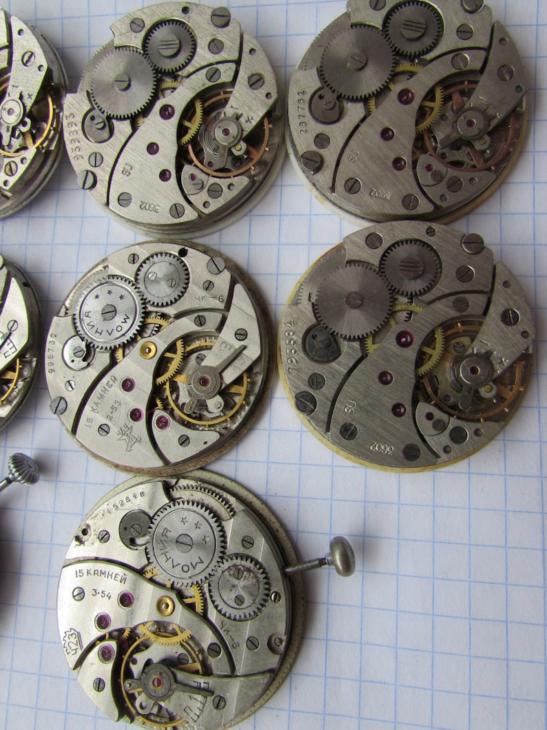 May include: Close-up of six antique watch movements with intricate gears and markings. The watch movements are silver and gold in color and have various numbers and text on them, including "15 KAMHEH", "2-53", "SU", "3602", "953393", and "237731".