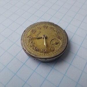 May include: A vintage pocket watch movement with a gold-toned face and silver-colored casing. The watch face displays Roman numerals and a smaller subdial. The hands point to approximately 5:20. The watch is aged and shows signs of wear.