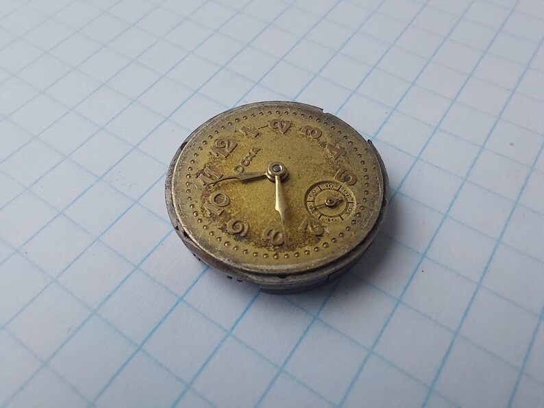 May include: A vintage pocket watch movement with a gold-toned face and silver-colored casing. The watch face displays Roman numerals and a smaller subdial. The hands point to approximately 5:20. The watch is aged and shows signs of wear.