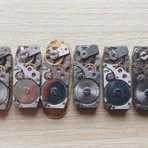 May include: A collection of vintage watch movements, showcasing intricate gears, jewels, and metal components. The mechanisms are arranged in a row, revealing the detailed inner workings of timepieces. The image highlights the craftsmanship and mechanical beauty of the watch parts.
