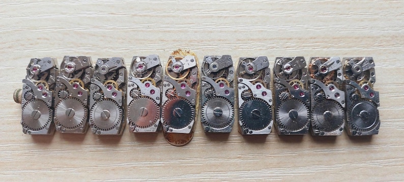 May include: A collection of vintage watch movements, showcasing intricate gears, jewels, and metal components. The mechanisms are arranged in a row, revealing the detailed inner workings of timepieces. The image highlights the craftsmanship and mechanical beauty of the watch parts.