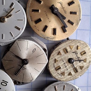 May include: A collection of vintage watch faces in various shapes and sizes. The faces are primarily silver and gold, with black hands and hour markers. Some faces have text, including "17 Rubis" and "Glash&uuml;tte".