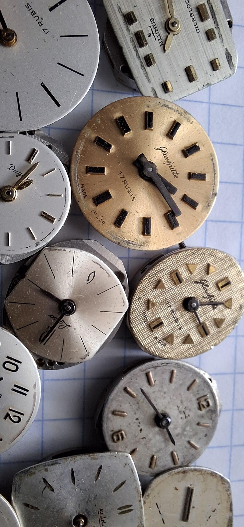 May include: A collection of vintage watch faces in various shapes and sizes. The faces are primarily silver and gold, with black hands and hour markers. Some faces have text, including "17 Rubis" and "Glash&uuml;tte".