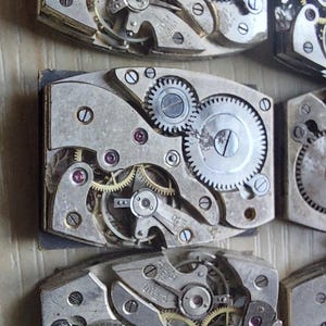 Peut inclure: Gros plan de plusieurs mouvements de montre vintage, pr&eacute;sentant des engrenages, des vis et des bijoux en rubis complexes. Les composants m&eacute;talliques argent&eacute;s sont dispos&eacute;s en formes rectangulaires, soulignant la pr&eacute;cision de l'horlogerie.
