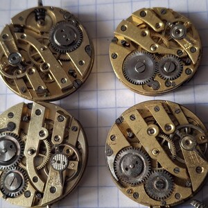 Op de afbeelding: Vier vintage uurwerkbewegingen met zichtbare tandwielen en ingewikkelde mechanische componenten. De cirkelvormige messing mechanismen hebben zilverkleurige tandwielen en schroeven, die de interne werking van antieke uurwerken laten zien. Deze horlogeonderdelen worden op een geruit oppervlak getoond.
