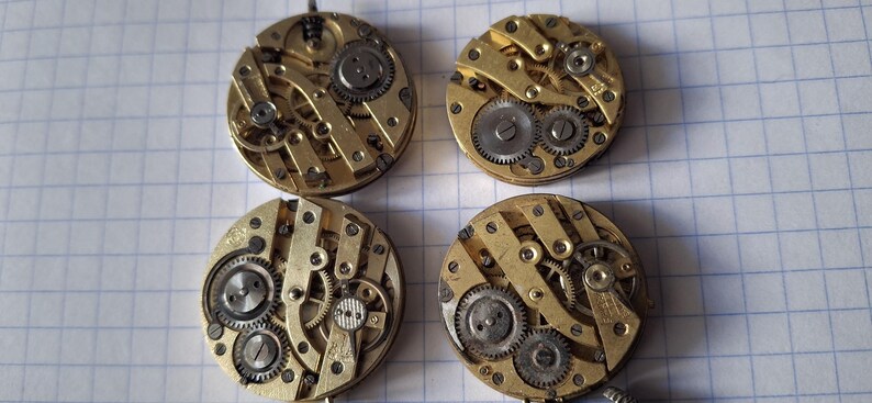 Op de afbeelding: Vier vintage uurwerkbewegingen met zichtbare tandwielen en ingewikkelde mechanische componenten. De cirkelvormige messing mechanismen hebben zilverkleurige tandwielen en schroeven, die de interne werking van antieke uurwerken laten zien. Deze horlogeonderdelen worden op een geruit oppervlak getoond.