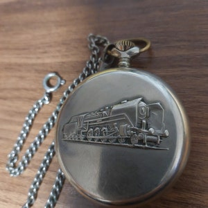 Puede incluir: Reloj de bolsillo plateado con un diseño de tren en relieve en la tapa. El reloj tiene una cadena plateada. El reloj está sobre un fondo de madera.