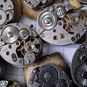 May include: Close-up of several disassembled watch movements, showcasing intricate gears, springs, and jewels. The metal components are silver and gold, with visible markings and text. The image highlights the mechanical complexity of vintage watchmaking.