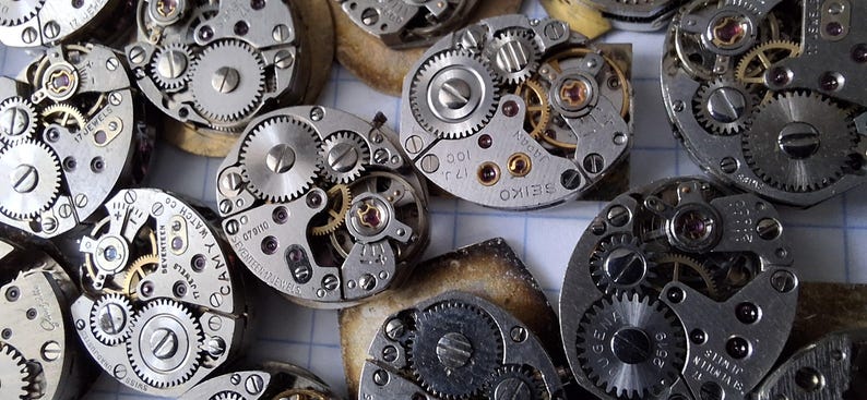 May include: Close-up of several disassembled watch movements, showcasing intricate gears, springs, and jewels. The metal components are silver and gold, with visible markings and text. The image highlights the mechanical complexity of vintage watchmaking.