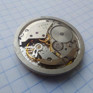 May include: Close-up of a vintage watch movement, showcasing intricate gears, springs, and other mechanical components. The silver-colored metal frame houses the internal mechanisms, with visible markings and numbers. The watch's internal workings are on display.