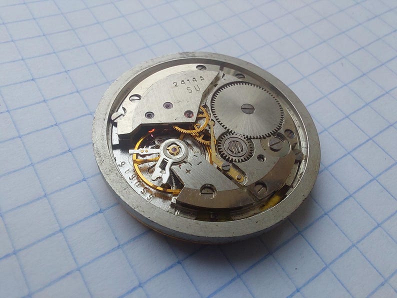 May include: Close-up of a vintage watch movement, showcasing intricate gears, springs, and other mechanical components. The silver-colored metal frame houses the internal mechanisms, with visible markings and numbers. The watch's internal workings are on display.
