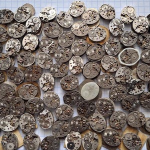 May include: A collection of vintage watch movements, showcasing intricate mechanical components. The silver-toned gears and plates are arranged on a white grid background, highlighting the detailed craftsmanship of each piece.
