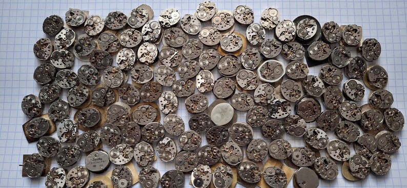 May include: A collection of vintage watch movements, showcasing intricate mechanical components. The silver-toned gears and plates are arranged on a white grid background, highlighting the detailed craftsmanship of each piece.