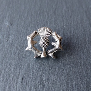May include: A silver-toned metal pin featuring a stylized thistle with a crown, surrounded by a wreath of thorns.