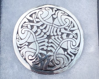 Pode incluir: Um broche redondo prateado com um intrincado design de nó celta, apresentado numa caixa de presente preta com acolchoamento branco. O broche apresenta padrões e curvas detalhadas, criando um efeito visual marcante. A caixa está parcialmente aberta.