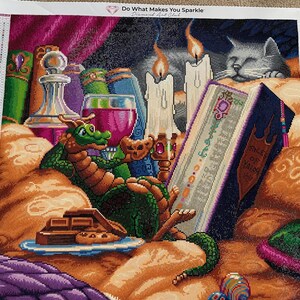 Könnte beinhalten: Ein Diamantgemälde mit einem grünen Drachen, Büchern, Kerzen und einer Katze. Der Drache liegt in der Nähe eines Buches mit dem Titel "Kings of Valor". Ein Glas Wein und ein Teller mit Schokolade sind ebenfalls zu sehen. Das Kunstwerk trägt den Titel "Do What Makes You Sparkle".