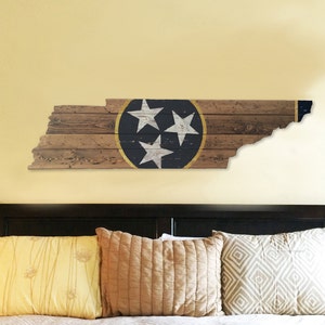 May include: A wooden wall art piece shaped like the state of Tennessee with the state flag painted on it. The flag features three white stars on a blue circle with a gold outline. The wood is painted in a distressed style with a brown and white color scheme.