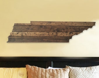 May include: A wooden wall art piece in the shape of the state of Tennessee. The art piece is made of reclaimed wood and has a rustic look. It is hanging on a yellow wall above a bed with brown bedding and pillows.