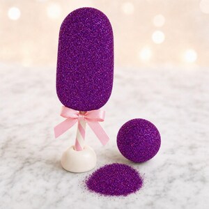 May include: A purple glitter-covered popsicle-shaped ornament on a white stick with a light pink ribbon bow, accompanied by a matching purple glitter sphere and a small pile of loose purple glitter on a white surface.