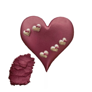 May include: A heart-shaped, burgundy-colored, decorative object with five gold hearts with pink bows attached to it. A small pile of burgundy-colored powder is in the lower left corner of the image.