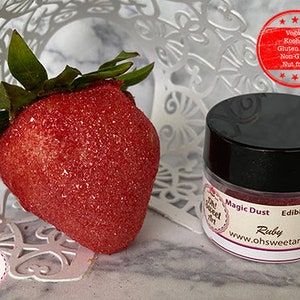 May include: A small jar of edible glitter labeled "Magic Dust Edible Glitter Ruby" with the website www.ohsweetart.com. The glitter is a vibrant red color. The jar is sitting on a white surface next to a red strawberry covered in red glitter.