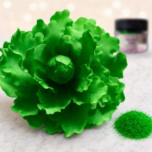 May include: A vibrant green sugar flower with ruffled, layered petals arranged in a dense, blooming peony shape. Beside the flower sits a small pile of bright green edible dust and a blurred black container with a label in the background.