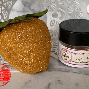 May include: A small jar of edible gold glitter labeled "Magic Dust Aztec Gold" by Oh! Sweet Art. The jar is sitting on a white surface next to a strawberry-shaped object covered in gold glitter.