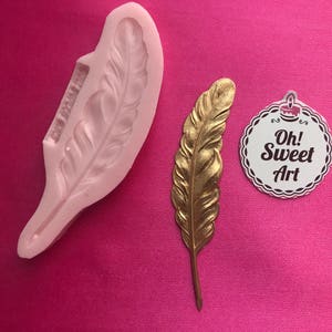 May include: A silicone mould shaped like a feather, with a gold-coloured feather next to it. The mould is white and the feather is shiny. The text "Oh! Sweet Art" is on a white circle sticker.