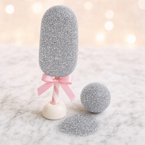 Pode incluir: Uma forma oval coberta de glitter prateado num pau às riscas com um laço de fita cor-de-rosa claro, assente numa pequena base branca. Ao lado encontra-se uma esfera de glitter prateado e um pequeno monte de glitter prateado.