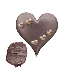 May include: A heart-shaped chocolate brown eyeshadow with a shimmery finish. The eyeshadow has five small gold hearts with bows on it.