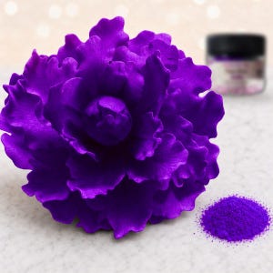 May include: A vibrant purple sculpted flower with layered, ruffled petals. Beside the flower sits a small pile of matching purple powder and a blurred black-lidded jar in the background, suggesting a craft or decorative supply set.
