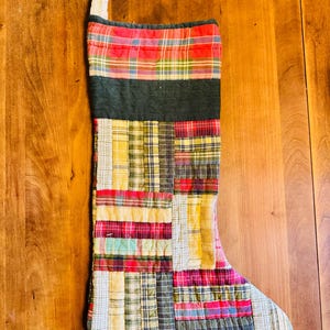 May include: A quilted Christmas stocking featuring a patchwork design of various plaid and striped fabrics in red, yellow, green, and white. The stocking has a solid dark green band near the top and a white fabric hanging loop at the corner.