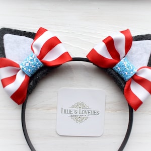 May include: A black headband featuring gray felt cat ears with white inner sections. Each ear is decorated with a red and white striped bow, centered with a blue glitter embellishment. The headband includes a tag with the text "Lillie's Lovelies".