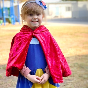 May include: A young child wearing a Snow White costume, featuring a blue and yellow dress and a red velvet cape. A white headband with a flower accent completes the look. The background is a park setting.