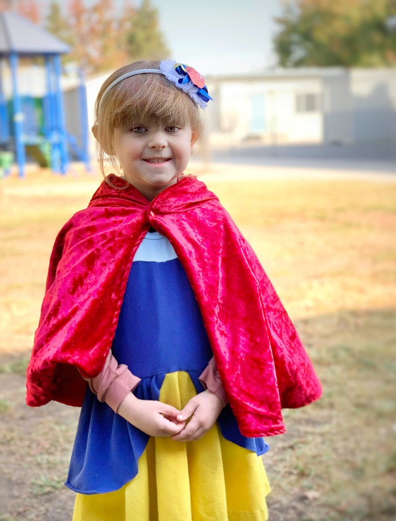 May include: A young child wearing a Snow White costume, featuring a blue and yellow dress and a red velvet cape. A white headband with a flower accent completes the look. The background is a park setting.