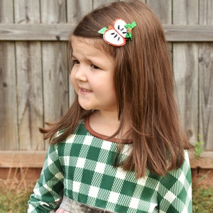 May include: A young girl with long brown hair wearing a green and white plaid dress with a pink skirt. She has an apple-shaped hair clip in her hair. The background is a wooden fence.