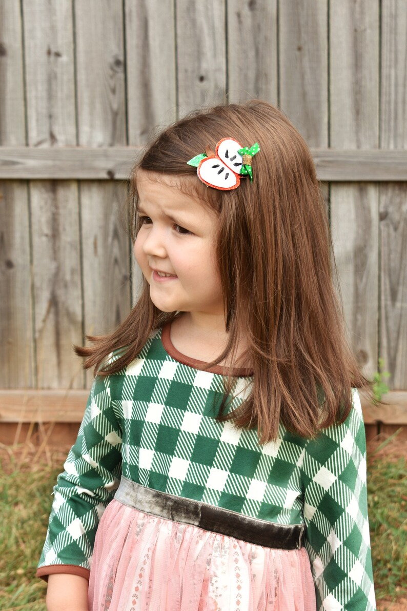 May include: A young girl with long brown hair wearing a green and white plaid dress with a pink skirt. She has an apple-shaped hair clip in her hair. The background is a wooden fence.
