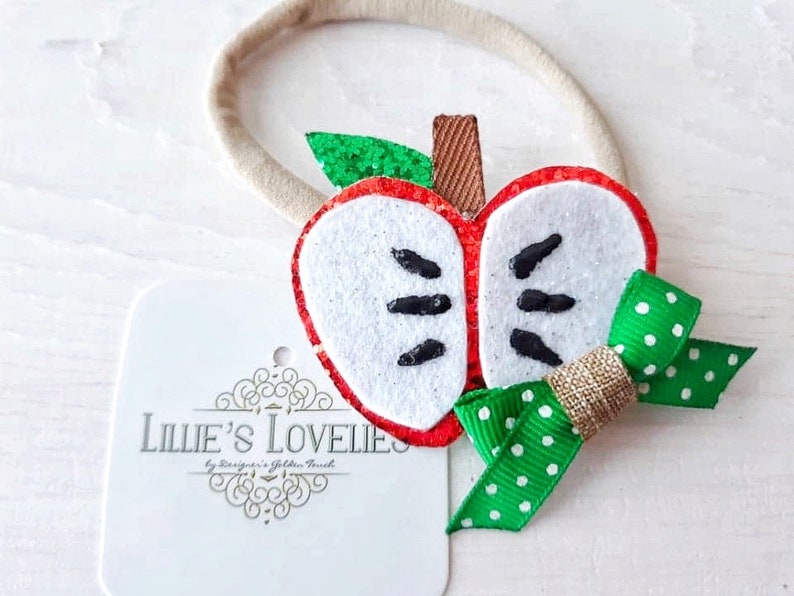 May include: A handmade apple-shaped baby headband. The apple is red with a white center, black seeds, and a green glitter leaf. A green polka dot bow and a gold ribbon accent the design. The headband is tan and the tag reads "Lillie's Lovelies".