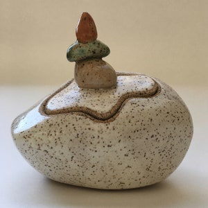 May include: A white ceramic box with a speckled finish, shaped like a rock. The lid has a small, stacked rock sculpture on top, with a brown, green, and orange glaze.