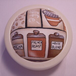 May include: A white ceramic pincushion with a round top and a fabric cover featuring a coffee theme. The fabric has a brown, beige, and white pattern with images of coffee beans, coffee cups, and coffee jars labelled "Mocha", "French Roast", and "House Blend".