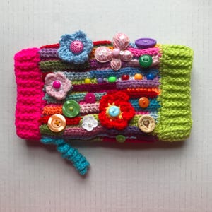 May include: A colorful, handmade crochet item with a variety of embellishments. The piece features a vibrant mix of colors, including pink, green, blue, and orange. Adorned with crochet flowers, buttons, and beads, it has a textured, tactile appearance.