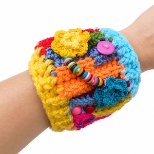 May include: A colorful, handmade crochet cuff bracelet. The bracelet features a patchwork design with vibrant hues of yellow, orange, red, blue, and pink. Embellishments include crochet flowers, buttons, and a beaded strand. The cuff is approximately 3 inches wide.