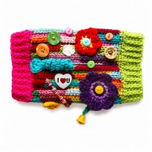 May include: A colorful, handmade crochet wristband featuring various embellishments. The wristband has a bright pink border and a green border. It is decorated with buttons, crocheted flowers, a bow, and a heart-shaped charm with the text "I U".